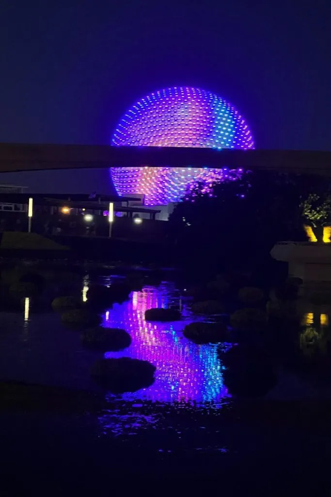 Photo of Spaceship Earth at night, lit up in purple, pink, turquoise, and blue, reflecting in the water in the foreground..