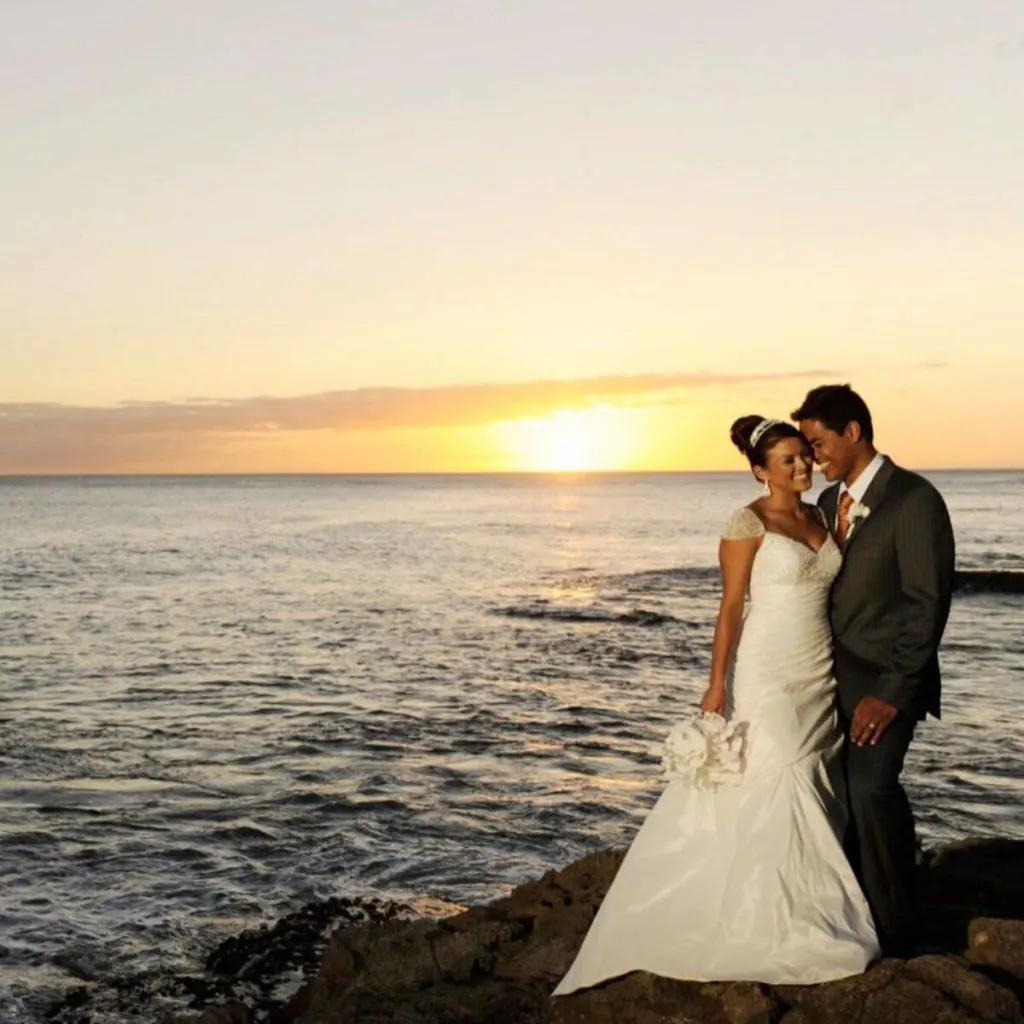 Photo of a bride a groom posing for a photo near the Aulani lagoon during sunset.