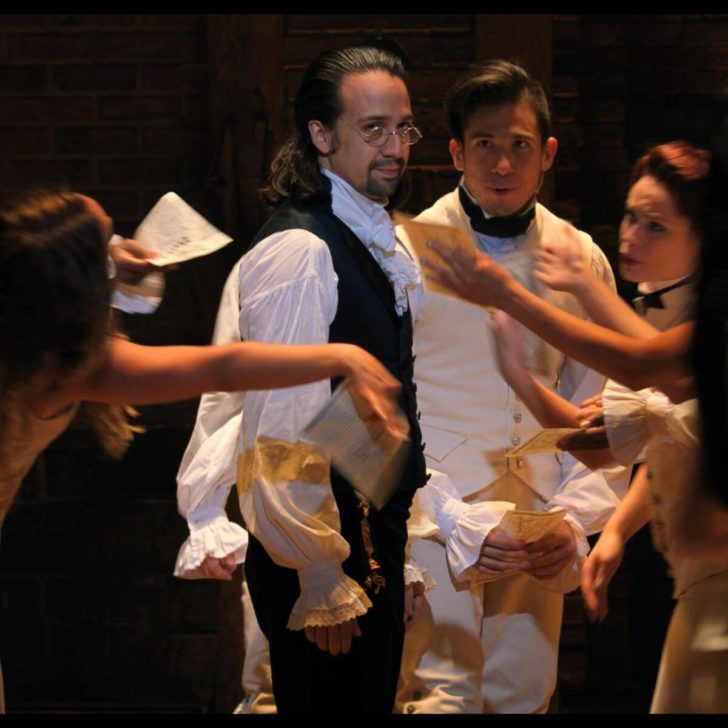 Closeup of Lin-Manuel Miranda as Alexander Hamilton in HAMILTON, the filmed version of the original Broadway production, with other cast members surrounding him.