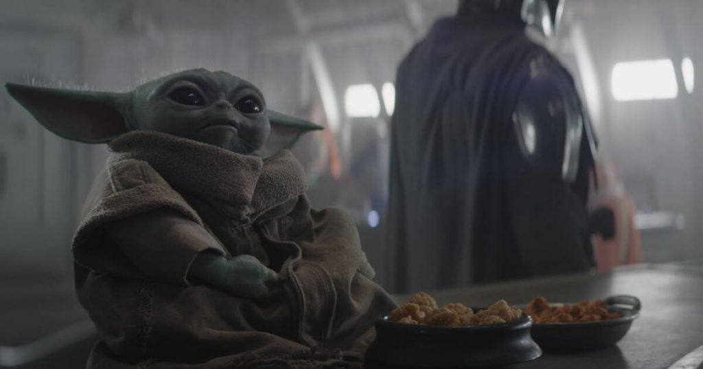 (L-R): Din Grogu sitting on a bar with bowls of food in front of him with Din Djarin (Pedro Pascal) in the background in Season 3 of Lucasfilm's The Mandalorian, one of the best shows to watch before going to Disney World.
