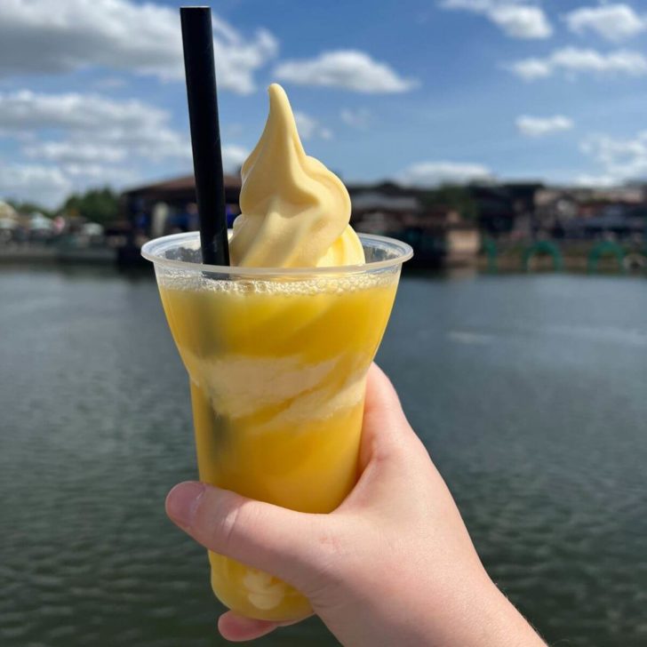 Photo of a hand holding a pineapple dole whip float with water in the background.