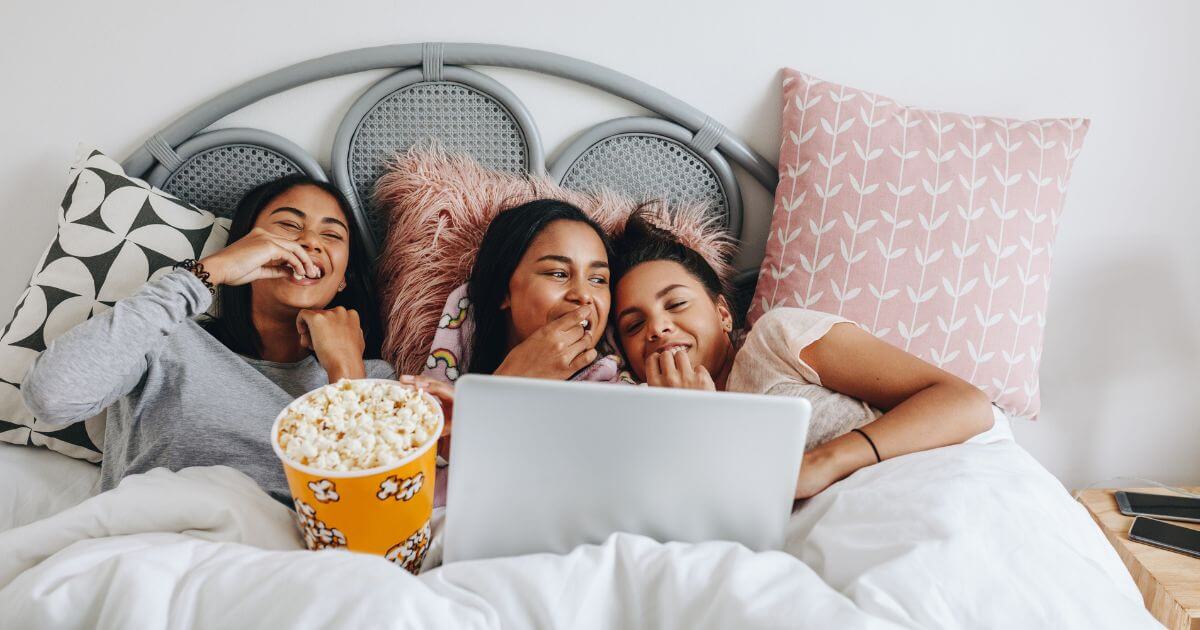 Photo of 3 young women relaxing on a bed while eating popcorn and watching a movie on a laptop.