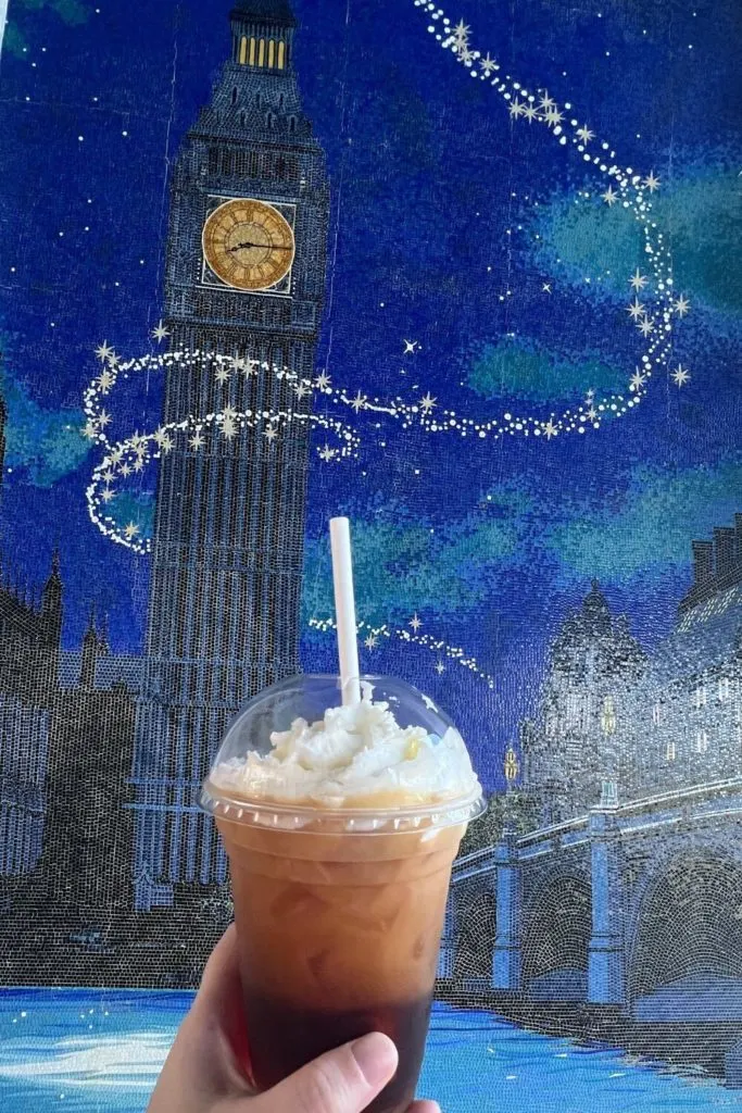 Photo of the Peter Pan in London mosaic at Disney's Riviera Resort with a person holding up a cinnamon cold brew coffee in front of it.