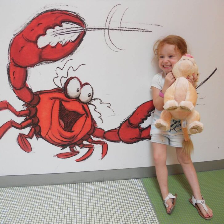 Photo of a young girl hugging a stuffed Nala toy, posing next to a drawing of Sebastian the crab on a wall at Disney's Art of Animation resort.