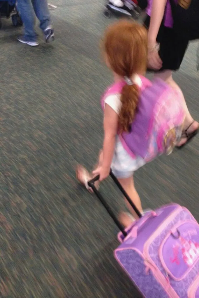 Photo of a young girl carrying a princess backpack and suitcase at an airport.