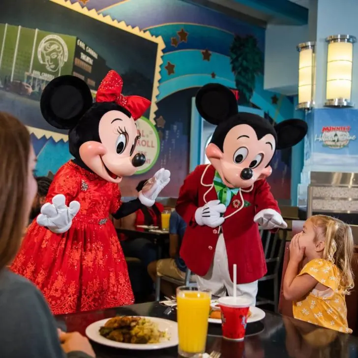 Photo of Minnie and Mickey Mouse dressed in their holiday finest, greeting a young girl at a dining table.