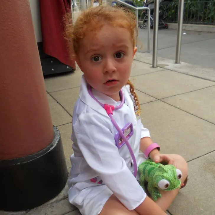 Photo of a toddler girl in a Doc McStuffins costume sitting on the ground while playing with a stuffed Pascal toy.