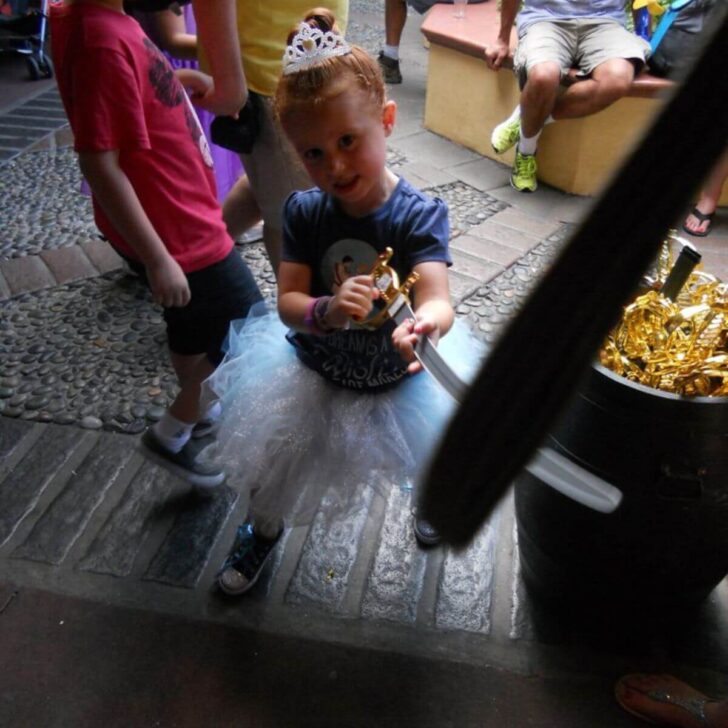 Photo of a young girl in a tiara and a tutu playing with a pirate sword outside the Pirates of the Caribbean ride at Magic Kingdom.