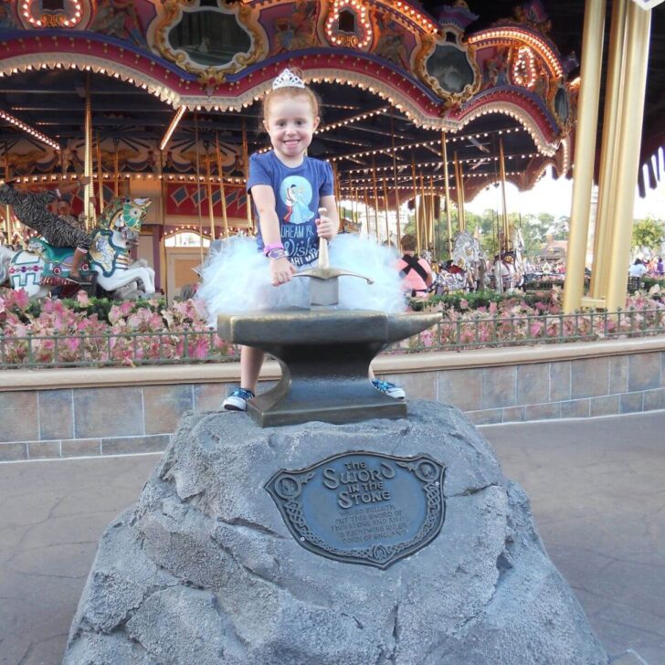 Photo of a girl in a tutu trying to pull the sword from the stone in front of the carousel at Magic Kingdom.