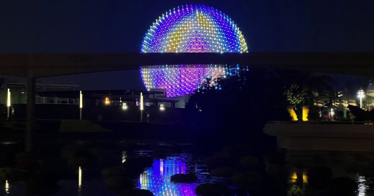 Photo of Spaceship Earth at Epcot at night, light up in iridescent colors while reflecting in water in the foreground.