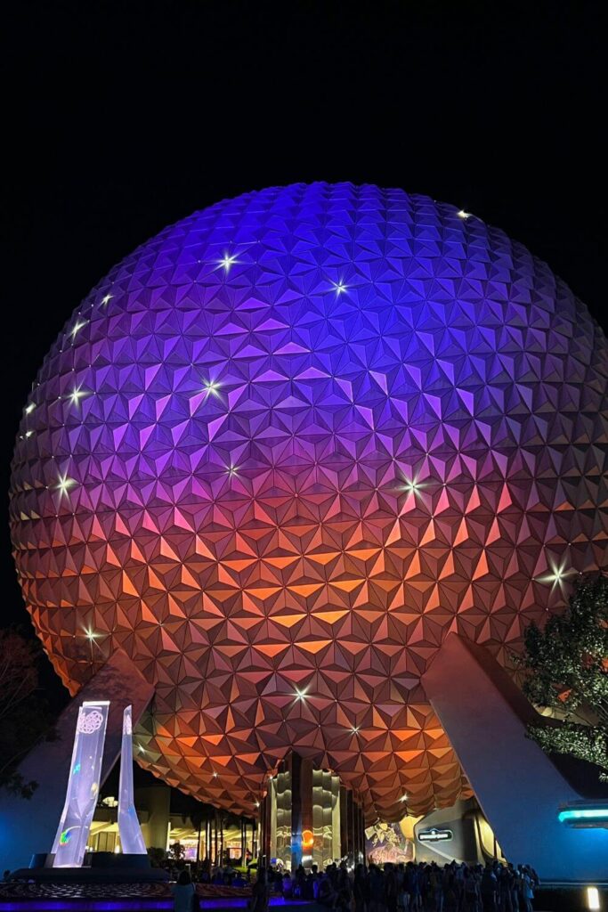 Photo of Spaceship Earth, aka the Epcot ball, lit up in blue, purple, pink, and orange with sparkles at night.