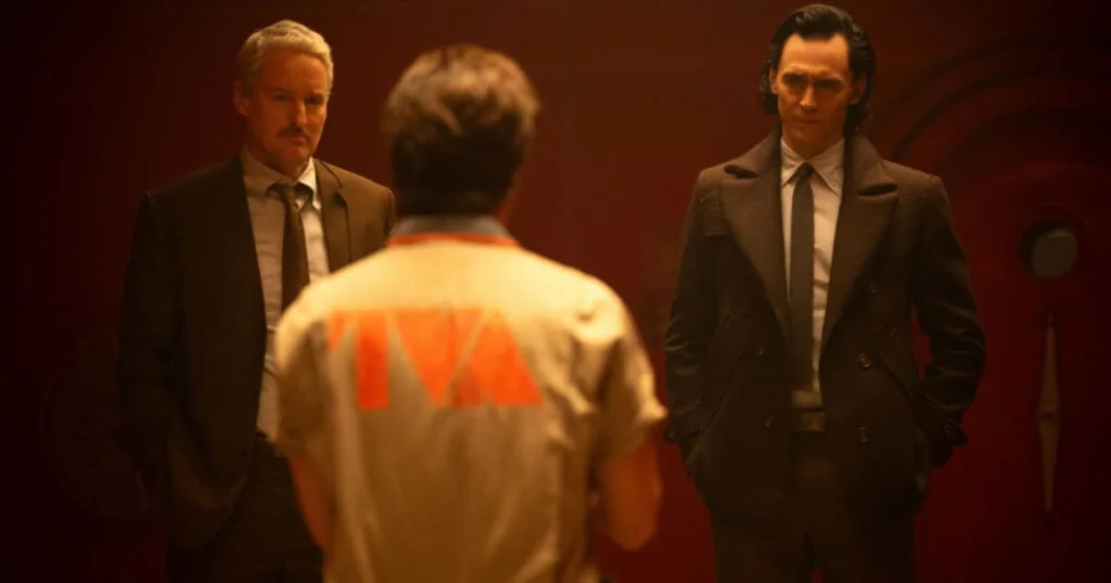 Promotional still featuring (L-R):Owen Wilson as Mobius, Rafael Casal as Hunter X-5, and Tom Hiddleston as Loki in Marvel Studios' LOKI, season 2, episode 2. Hunter X-5 has his back to us, showing an orange TVA logo on a khaki prisoner jumpsuit. Mobius and Loki are looking at him with serious expressions on their face while wearing their business best.