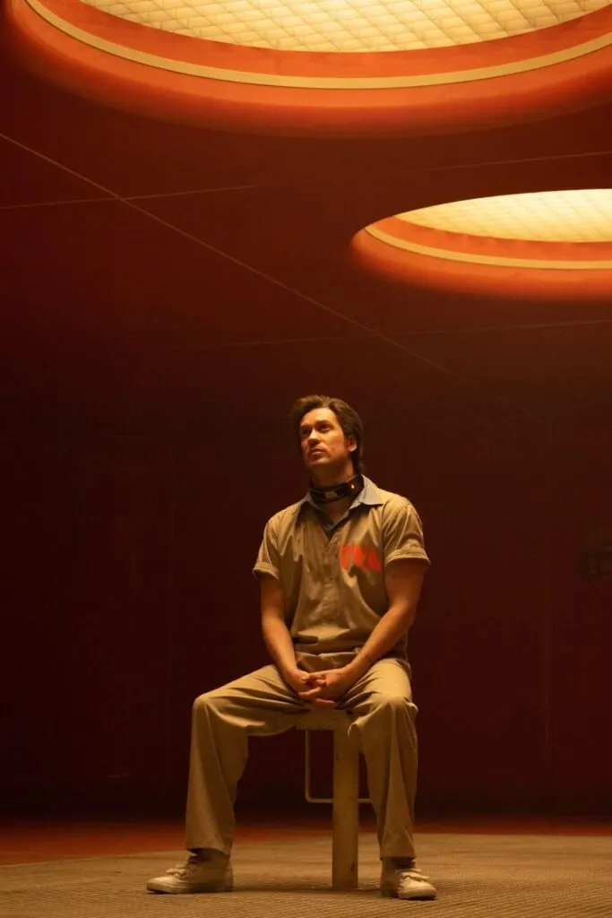 Photo still featuring Rafael Casal as Hunter X-5 as a TVA prisoner in Marvel Studios' LOKI, season 2, episode 2. He is sitting on a stool under bright orange lights, wearing a khaki TVA prisoner jumpsuit and time collar.