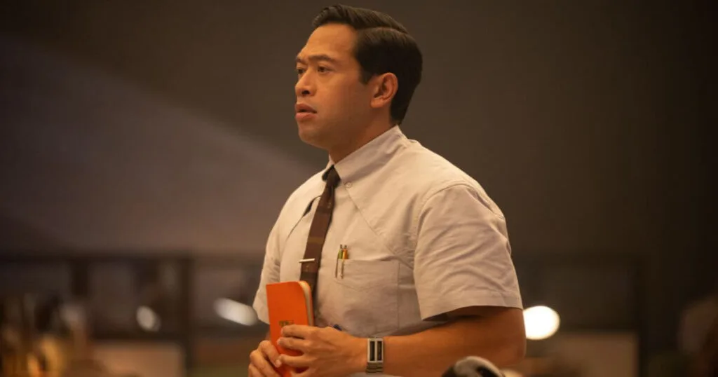 Photo featuring Eugene Cordero as Casey in Marvel Studios' LOKI, Season 2. He is holding an orange TVA guidebook in his hands with a concerned look on his face.