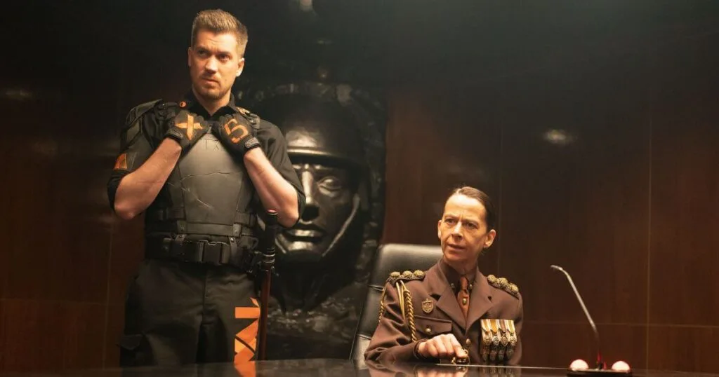 Photo still featuring (L-R): Rafael Casal as Hunter X-5 and Kate Dickie as General Dox in Marvel Studios' LOKI, Season 2, Episode 1. General Dox is sitting at the head of the table, with a stern look, pointing her finger. Hunter X-5 stands to her right in a defensive pose.