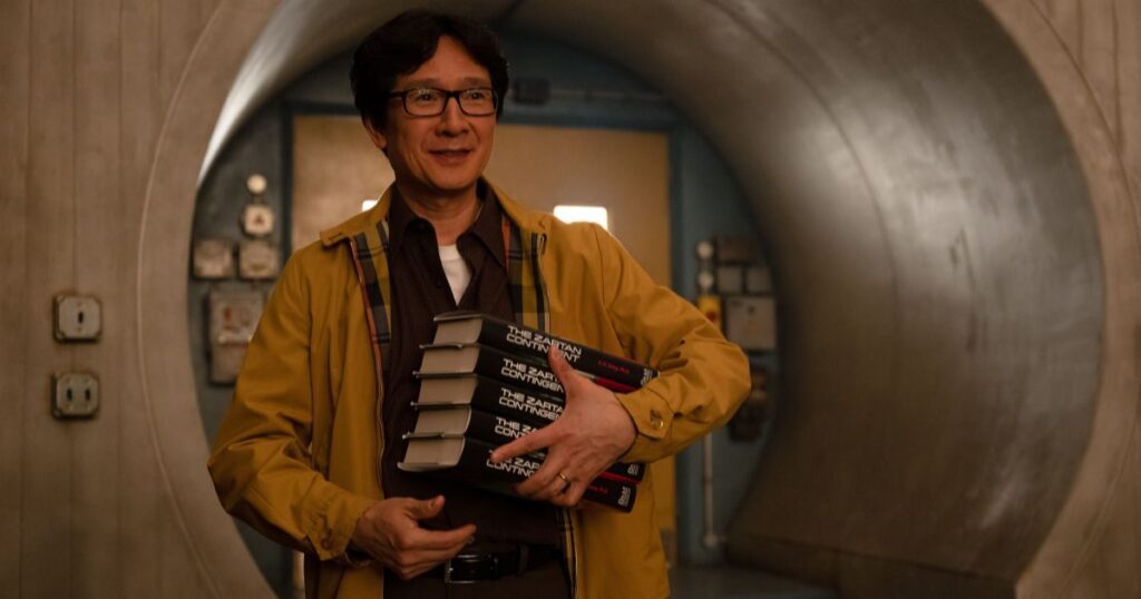 Photo still of Ke Huy Quan as A.D. Doug, aka O.B., in Marvel Studios' LOKI, Season 2, episode 5, carrying a stack of books called The Zartan Contingent.