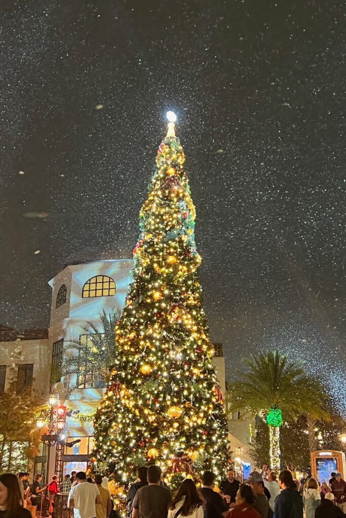 Photo of a tall Christmas tree lit up at night with white lights while faux snow falls at Disney Springs Town Center.