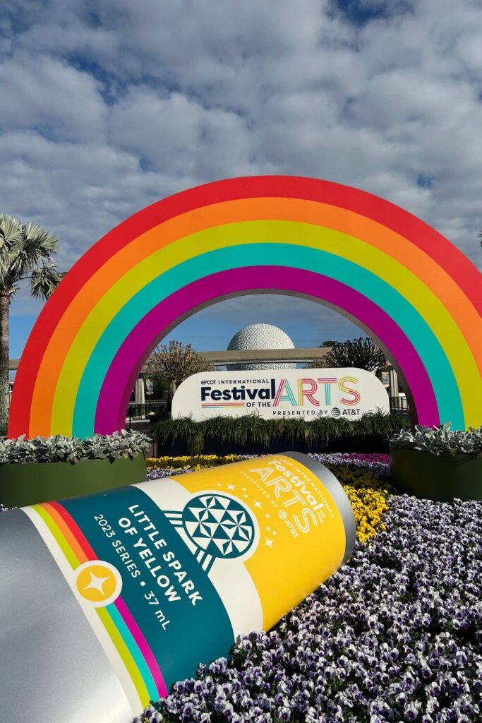 Vertical photo of a display for the Epcot Festival of the Arts with a large rainbow and paint tube, with Spaceship Earth in the background.