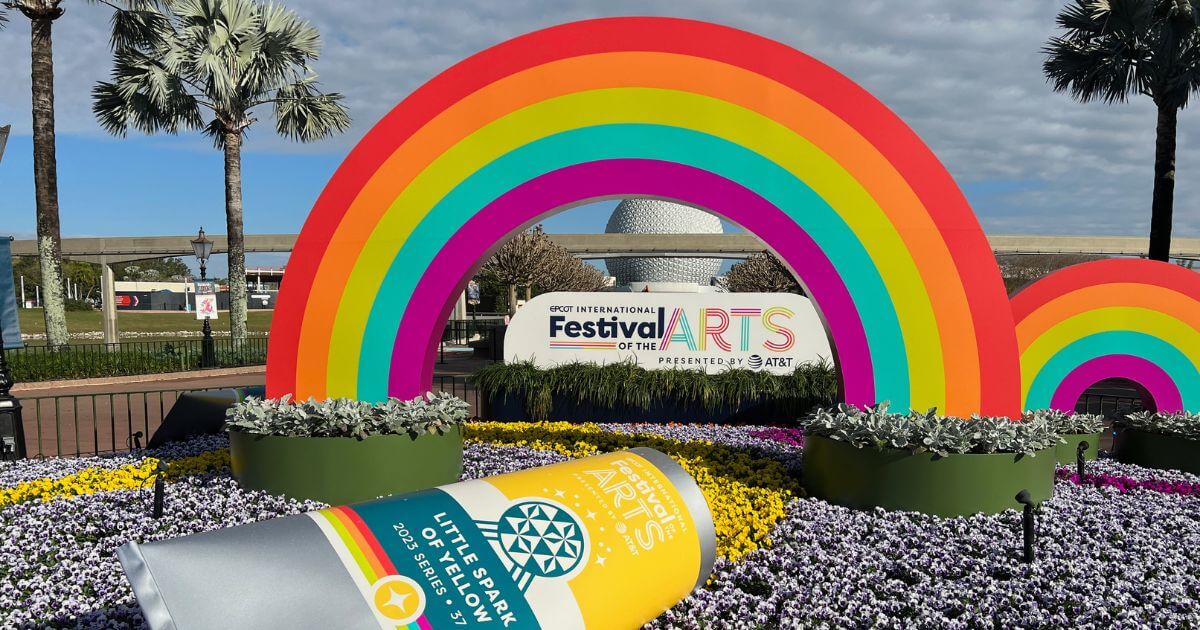 Horizontal photo of a floral and art display for the Epcot Festival of the Arts with a large rainbow and paint tube, with Spaceship Earth in the background.