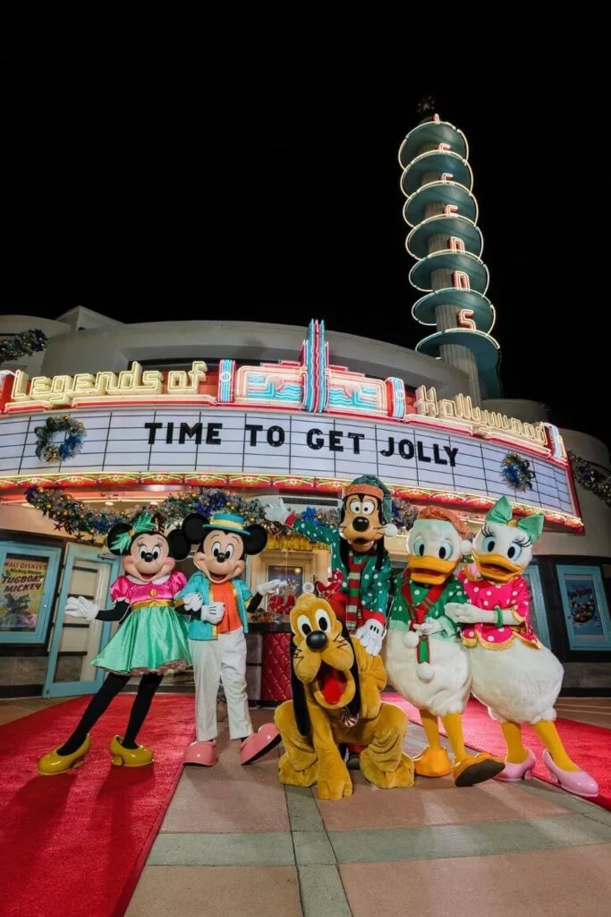 Photo of (L-R) Minnie Mouse, Mickey Mouse, Pluto, Goofy, Donald Duck, and Daisy Duck all in holiday inspired outfits, posing outside of Legends of Hollywood at Hollywood Studios. A sign above them on the marquee reads: Time to Get Jolly.