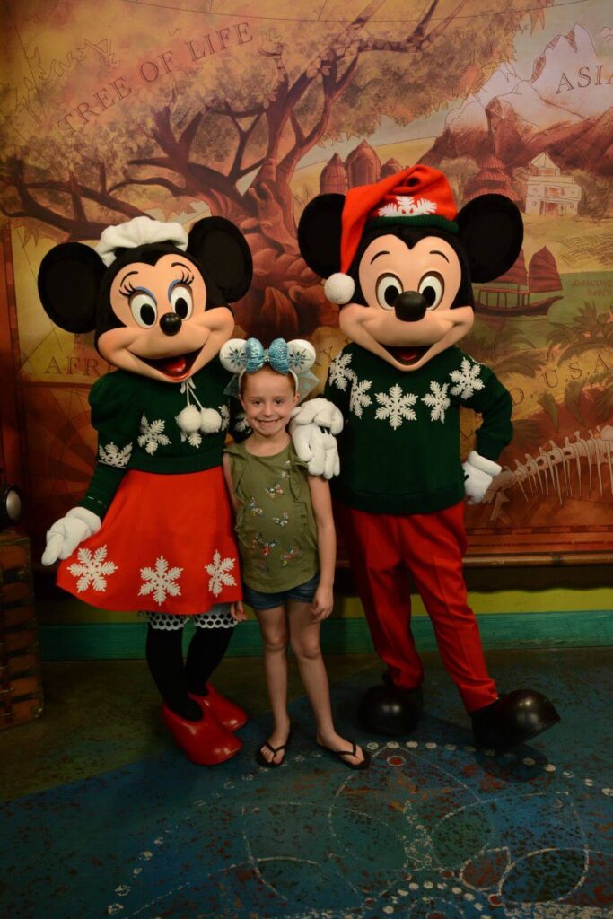 Photo of (L-R) Minnie Mouse in holiday outfit, a young girl with Frozen themed ears, and Mickey Mouse in holiday outfit and Santa hat.