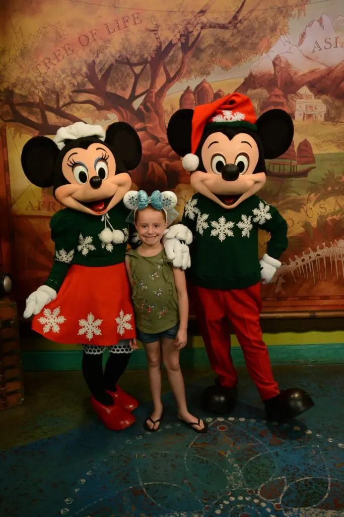 Photo of (L-R) Minnie Mouse in holiday outfit, a young girl with Frozen themed ears, and Mickey Mouse in holiday outfit and Santa hat.