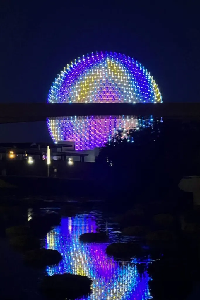 Photo of Spaceship Earth with holographic lights at night, reflecting in water in the foreground.