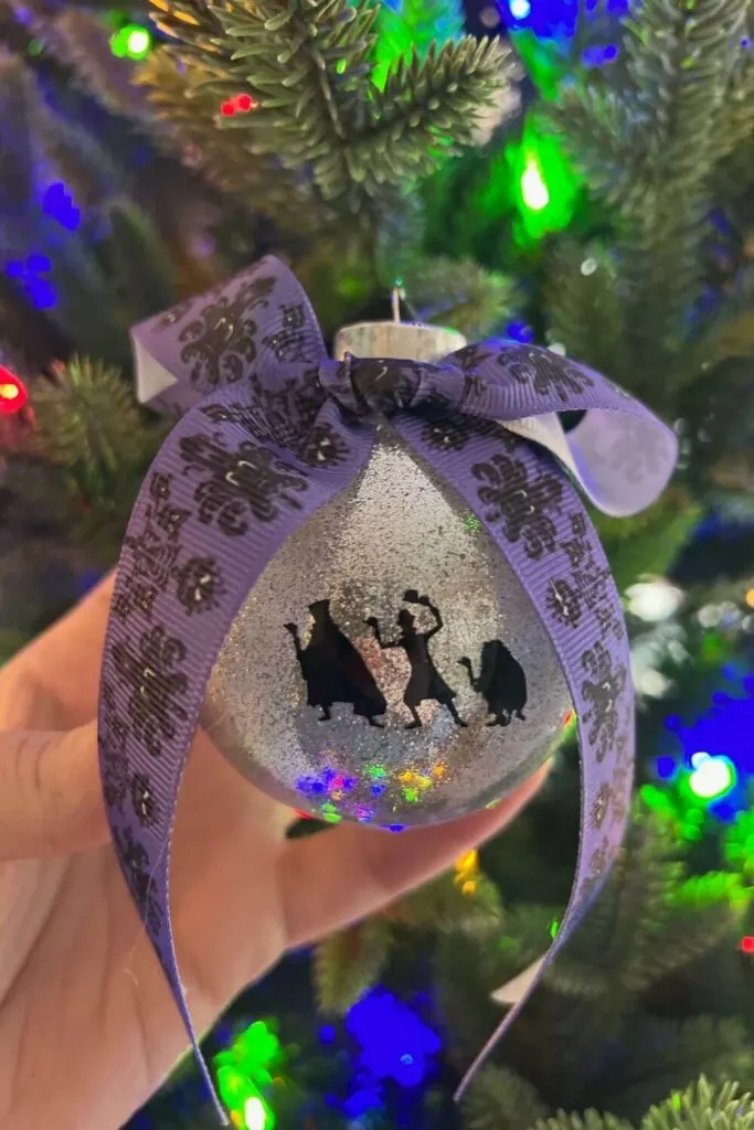 Closeup of a DIY Haunted Mansion ornament with a silver glitter base, a hitchhiking ghosts sticker, and a purple Haunted Mansion bow.