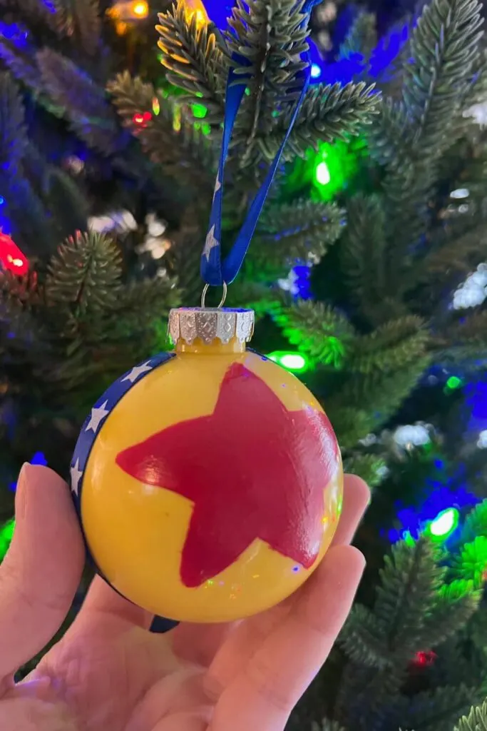 Closeup of a DIY ornament that looks like the Pixar ball. It has a yellow base with a red star in the middle and a blue ribbon with white stars wrapped around it vertically.