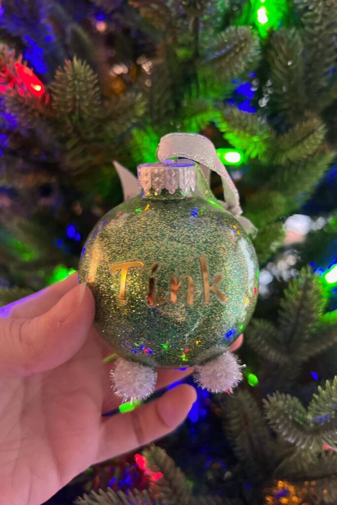 Closeup of a lime green glitter ornament with white and silver pom poms for feet and silver wings. In the middle are gold stickers that spell out: Tink.