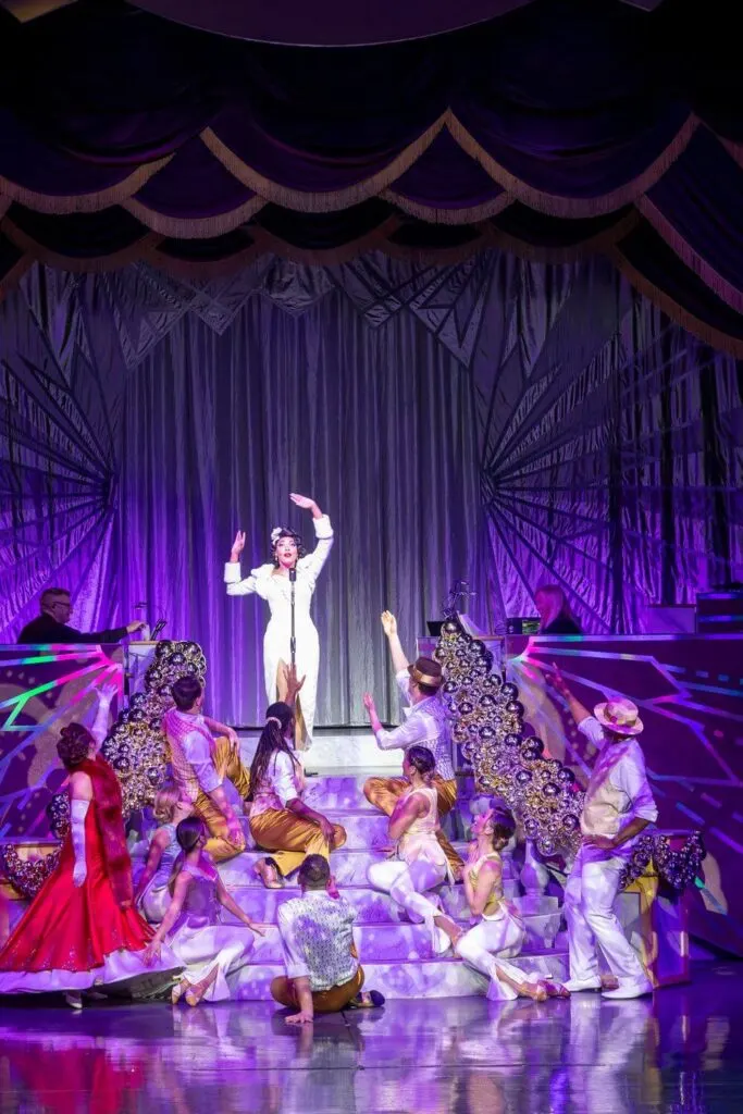 Photo of Tiana performing in the variety show at Disney Jollywood Nights, with other performers watching her from steps in the foreground.