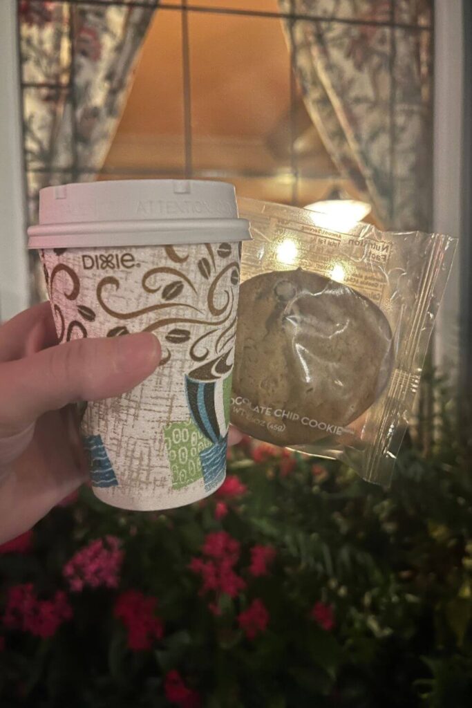 Closeup of a cup of hot cocoa and a prepackaged chocolate chip cookie with a restaurant window in the background.