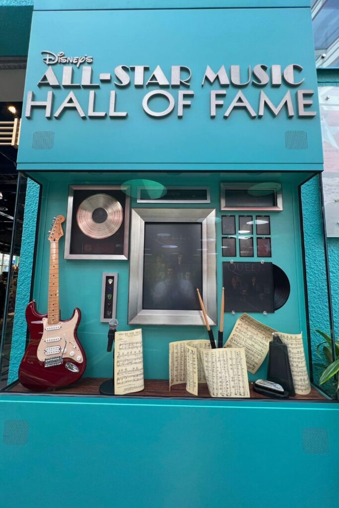 Photo of a museum-like display featuring photos, instruments, and records of the rock band, Queen. Text on the display reads: Disney's All-Star Music Hall of Fame.