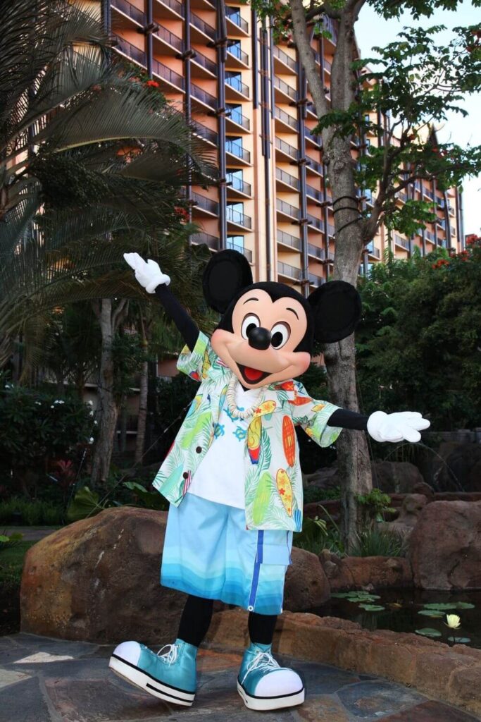 Photo of Mickey Mouse at Aulani, wearing an Aloha shirt, puka shell necklace, and board shorts.