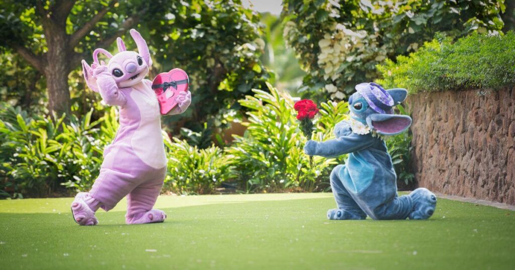 Photo of Stitch on one knee, holding flowers out at Angel, who's holding a heart-shaped box of chocolates while posing shyly.