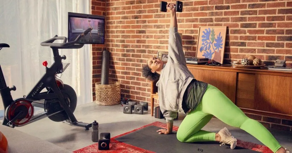 Photo of a woman doing a strength workout with a Peloton bike in the background.