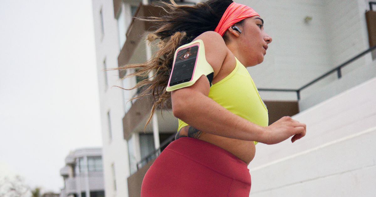Photo of a woman running outdoors with her phone on her upper arm, open to the Peloton app.