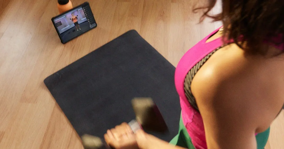 Photo looking over a woman's shoulder as she works out at home with the Peloton app playing a workout on her iPad in the background.