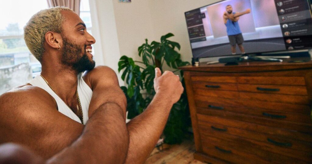 Photo of a young man stretching with the Peloton app playing a workout on a television in the background.