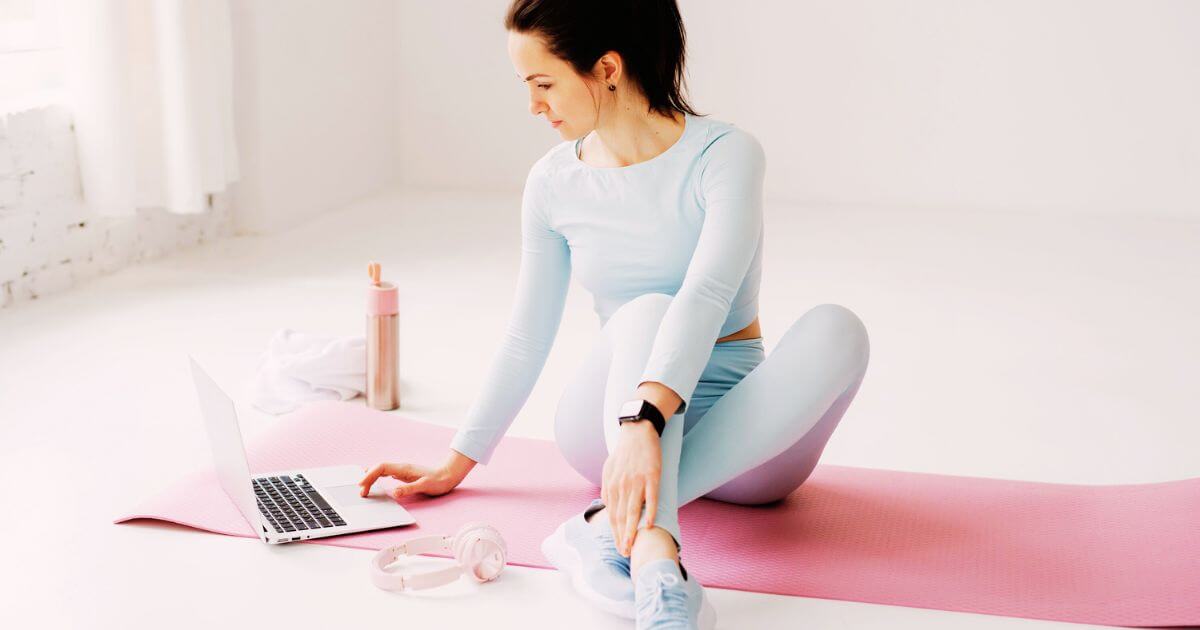 Photo of a woman in workout gear sitting on a yoga mat while setting up an at-home class on her laptop.