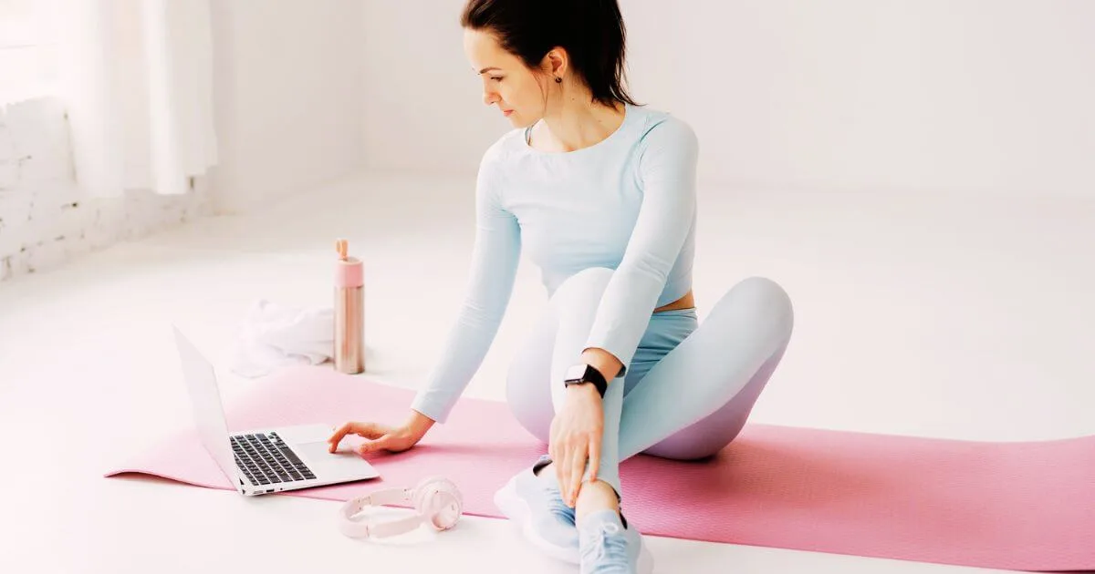 Photo of a woman in workout gear sitting on a yoga mat while setting up an at-home class on her laptop.