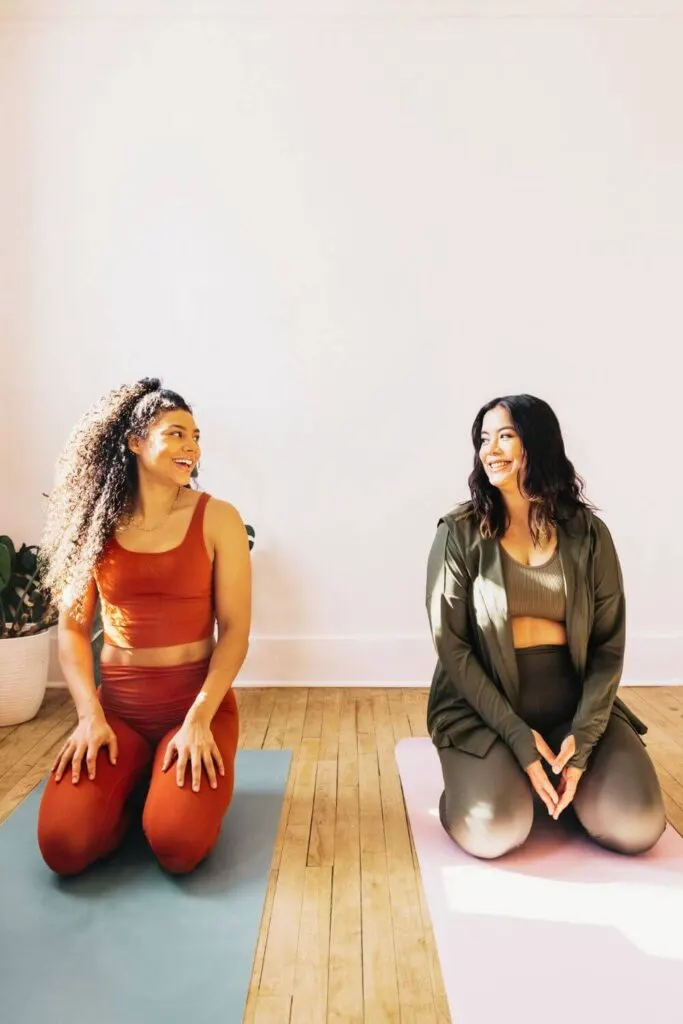 Photo of two women in workout gear, kneeling on yoga mats, smiling at each other.