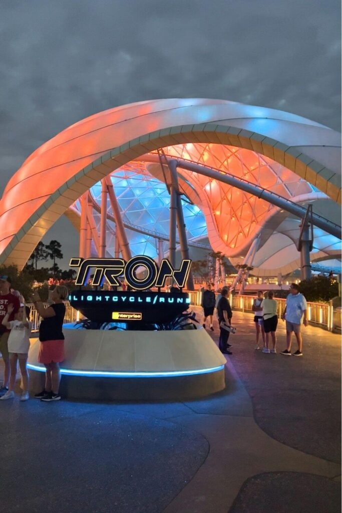 Vertical photo of the entrance to TRON Lightcycle Run at Disney World's Magic Kingdom. The canopy above the roller coaster is a mix of neon blue and orange lights with cloudy skies in the background.