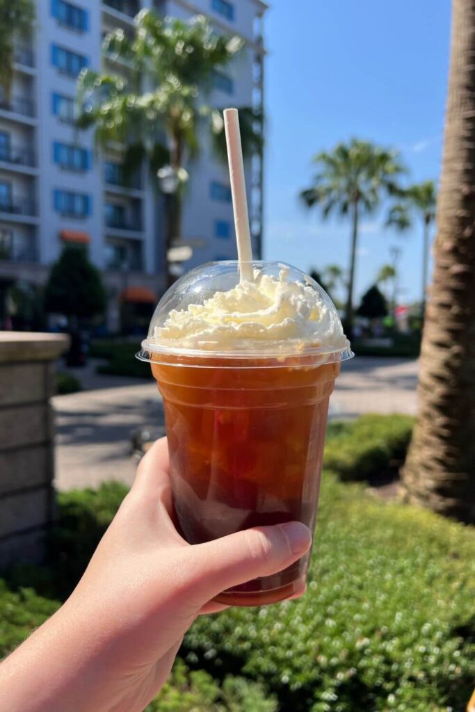 Photo of a person holding the cinnamon iced coffee from Disney's Riviera Resort with the resort in the background.