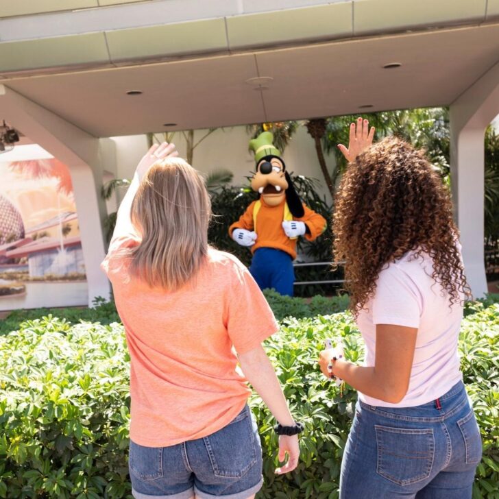 Photo from behind two women in t-shirts, waving at Goofy in the background.