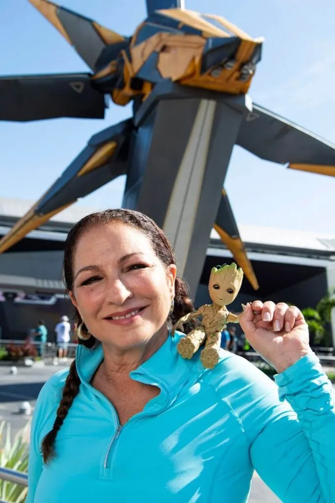 Photo of singer Gloria Estefan with a tiny plush Groot on her shoulder posing in the courtyard for Cosmic Rewind with the starcruiser in the background.