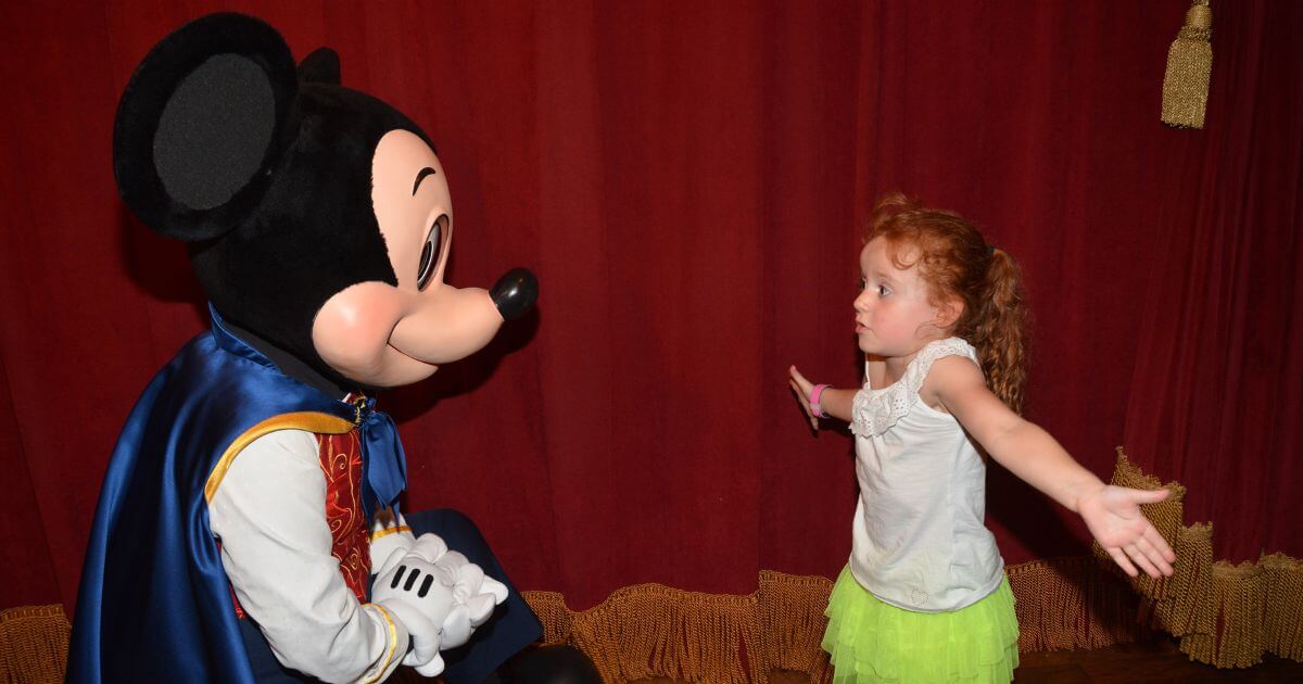 Photo of a young girl shrugging her shoulders as she talks to Mickey Mouse.