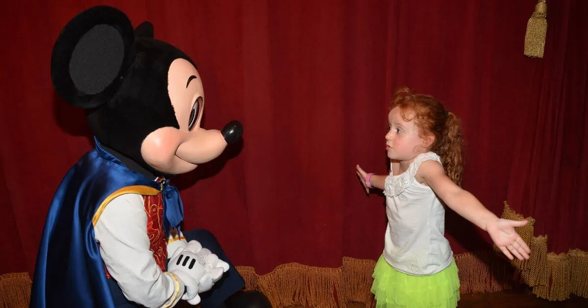 Photo of a young girl shrugging her shoulders as she talks to Mickey Mouse.