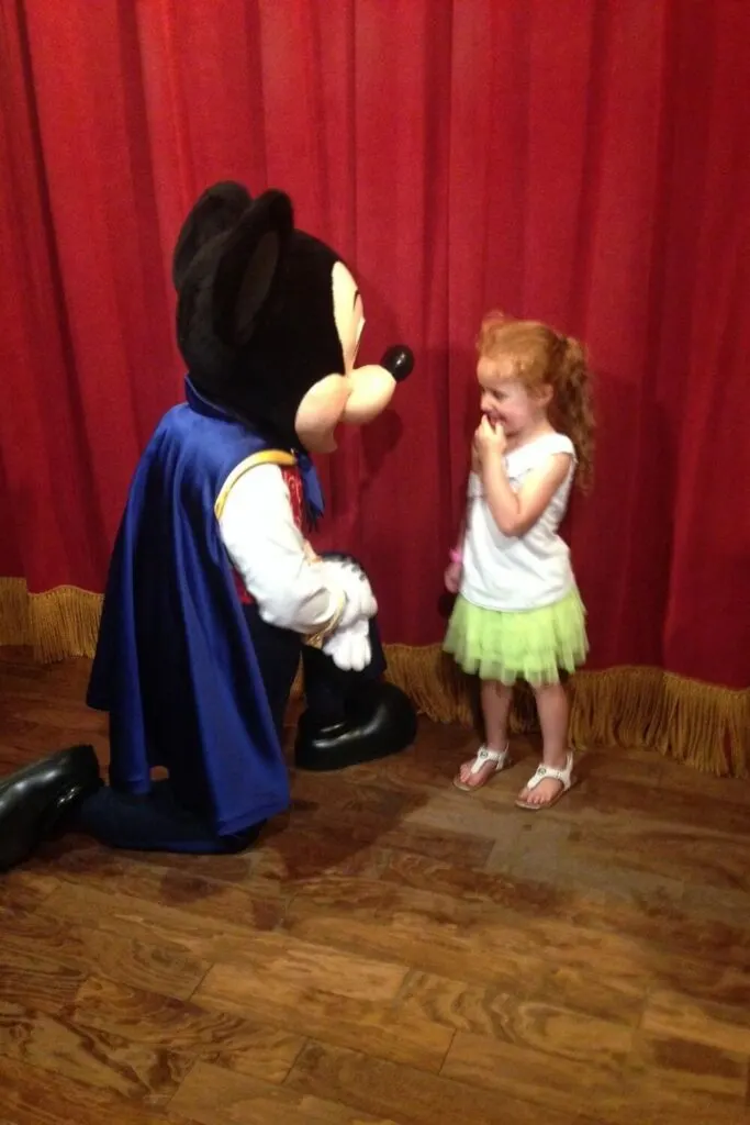 Photo of a young girl chatting with Mickey Mouse, who is down on one knee to meet her height.