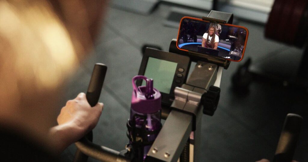 Promotional photo of a person on an exercise bike with their mobile phone open to the Peloton app.